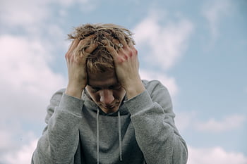 Young man in gray hoodie holds head in frustration, set against cloudy sky. HD wallpaper