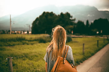 A woman with blonde hair and a backpack explores a scenic mountain landscape at sunset, capturing a sense of dom and travel. HD wallpaper