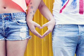 Close-up of a young couple forming a heart shape with their hands against a vibrant yellow background. HD wallpaper