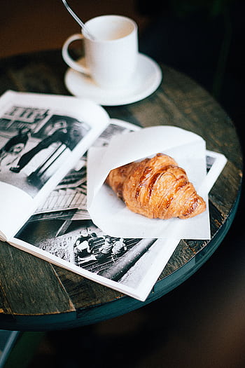 A cozy setup featuring a warm croissant and coffee cup on a wooden table with an open magazine. HD phone wallpaper