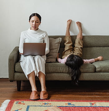 Mother multitasking with laptop while daughter plays on sofa, balancing work and family. HD phone wallpaper