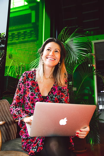 Smiling woman using a laptop in a modern indoor setting with lush greenery. HD phone wallpaper