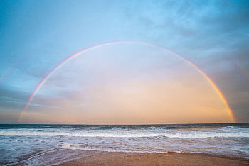 A stunning rainbow arches over a serene beachfront at sunset, creating a tranquil seaside atmosphere. HD wallpaper