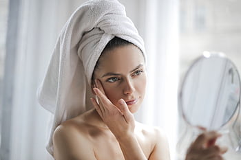 A young woman wrapped in a towel examines her skin in a mirror, focusing on skincare and relaxation. HD wallpaper
