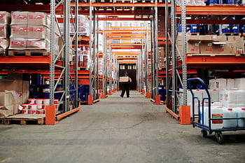 A worker carrying a box in a well-organized warehouse storage aisle. HD wallpaper