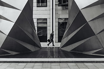 Monochrome of a businessman walking between geometric sculptures in a city setting. HD wallpaper