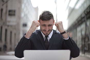 A joyful businessman in a suit triumphantly celebrates success on his laptop outdoors. HD wallpaper