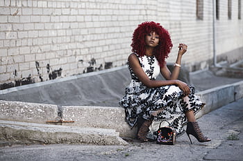 Stylish young woman with curly red hair and patterned dress sitting by a brick wall. HD wallpaper