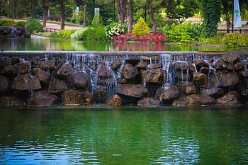 A serene waterfall over rocks in Şehitkamil Park, Gaziantep, surrounded by lush greenery and flowers. HD wallpaper