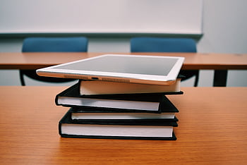 A tablet rests on top of a stack of books in an empty classroom, illustrating modern education. HD wallpaper