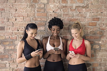 Three diverse women smiling and using smarts indoors against a brick wall. HD wallpaper