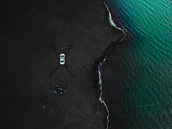 Drone shot capturing a car on a black sand beach by teal ocean waves. HD wallpaper
