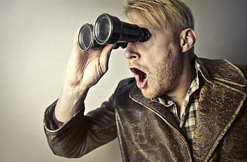 A surprised man in a leather jacket peers through binoculars with a shocked expression. HD wallpaper