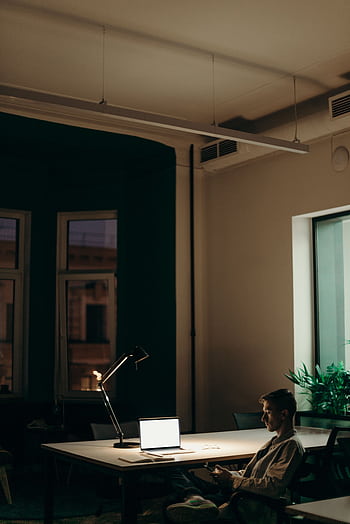 Man working late in an office using a laptop under a desk lamp, showcasing dedication and focus. HD phone wallpaper