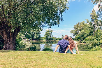 A couple sitting by a lake enjoying a sunny day in a lush park. HD wallpaper