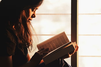 A woman engrossed in reading a book by a sunlit window creates a serene atmosphere. HD wallpaper