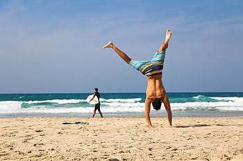 Man doing a handstand on a sunny beach with surfer in background, evoking a sense of dom and joy. HD wallpaper