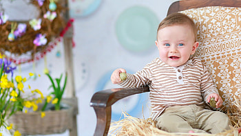 Smiling Blue Eyes Cute Baby Boy Is Sitting On Chair Wearing Brown White Lines Top And Pant In Blur Background Cute HD wallpaper