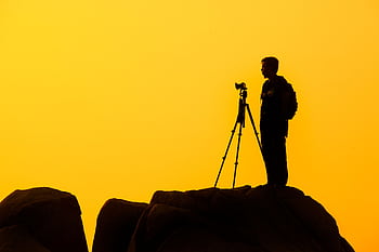 Silhouette of a lone grapher with tripod on a rocky cliff against a golden sunset sky. HD wallpaper