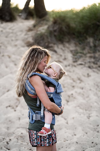 A loving mother kisses her toddler in a baby carrier on a sandy beach, capturing a tender parenting moment. HD phone wallpaper