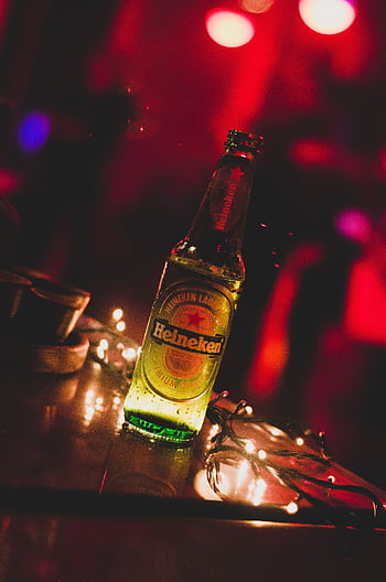 Close-up of a Heineken beer bottle illuminated by string lights in a vibrant party setting. HD phone wallpaper