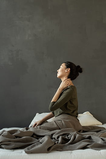 Side view of a woman sitting on a bed indoors, massaging her neck for stress relief. HD phone wallpaper