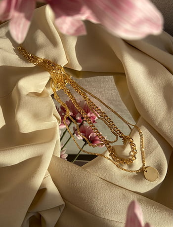 Gold necklaces lie elegantly on beige fabric with pink flowers in a sunlit setting. HD phone wallpaper
