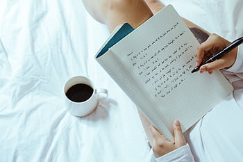 An individual writing in a notebook with a cup of coffee nearby, set against a cozy indoor background. HD wallpaper