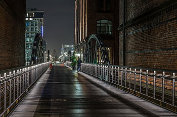 Modern urban bridge lit at night with buildings and a person walking, creating a serene city ambiance. HD wallpaper