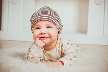 A smiling baby wearing a striped beanie and cozy outfit lies on a soft carpet indoors. HD wallpaper