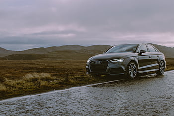 A luxury Audi sedan parked on a wet mountain road with moody skies and a scenic backdrop. HD wallpaper