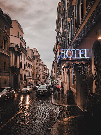 A rainy city street scene at dusk, featuring a glowing neon hotel sign, wet cobblestones, and bustling cars. HD phone wallpaper