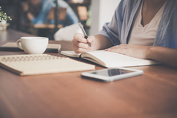 A woman writes in a notebook at a café table with a coffee and smart nearby. HD wallpaper