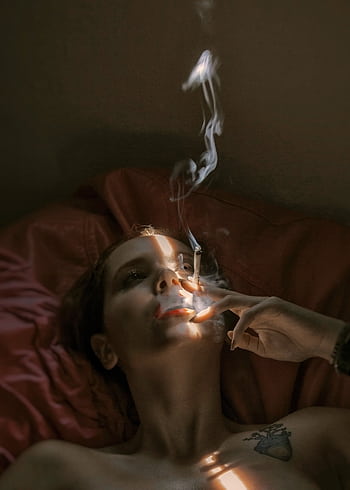 A woman lying on a bed indoors, smoking a cigarette with dramatic lighting casting shadows on her face. HD phone wallpaper