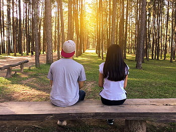 A young couple sits on a bench surrounded by tall trees in a forest at sunset. HD wallpaper