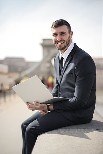 Happy businessman in suit holding laptop while sitting on a city bridge, showcasing modern corporate lifestyle. HD phone wallpaper