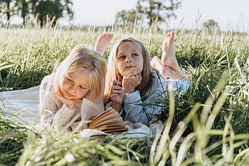 Two young girls lying on a picnic blanket in a sunny field, reading and daydreaming. HD wallpaper