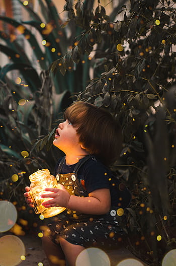 A young child gazes in wonder while holding a jar of lights in a magical forest setting, capturing innocence and curiosity. HD phone wallpaper
