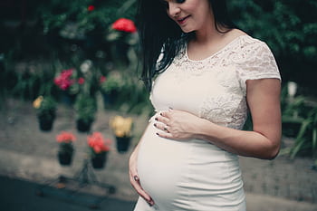 Pregnant woman in white lace dress, posing outdoors with flowers, exuding happiness. HD wallpaper