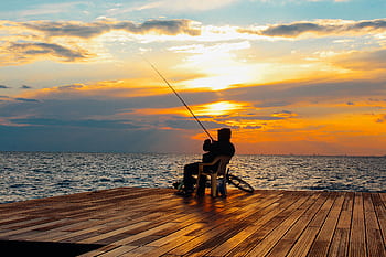 A fisherman sits on a wooden deck, enjoying a peaceful sunset by the ocean, rod in hand. HD wallpaper