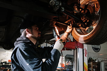 Mechanic examining car's undercarriage at a garage, focusing on vehicle maintenance. HD wallpaper