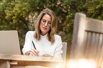 A woman in glasses writing and working on her laptop outdoors in natural daylight. HD wallpaper