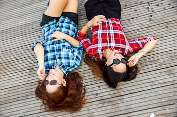 Two women in plaid shirts lying on a wooden deck, enjoying a sunny day with sunglasses. HD wallpaper