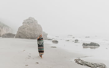 A serene scene of a woman enjoying a solitary walk on a misty beach with rocky coastal views. HD wallpaper