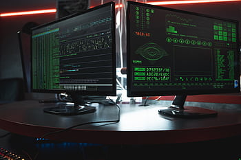 Close-up of dual computer monitors with green coding interfaces in a dark room, highlighting cyber security themes. HD wallpaper