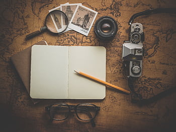 A flat lay of travel essentials on a vintage map surface, including a camera, notebook, and magnifying glass. HD wallpaper