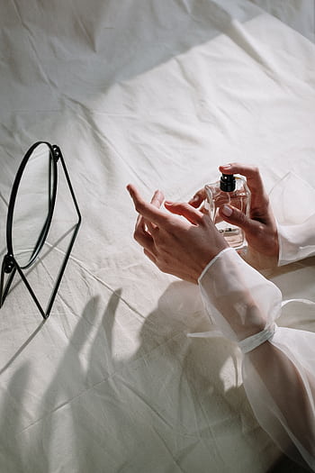 Delicate hands holding a perfume bottle beside a mirror on a white cloth background. HD phone wallpaper