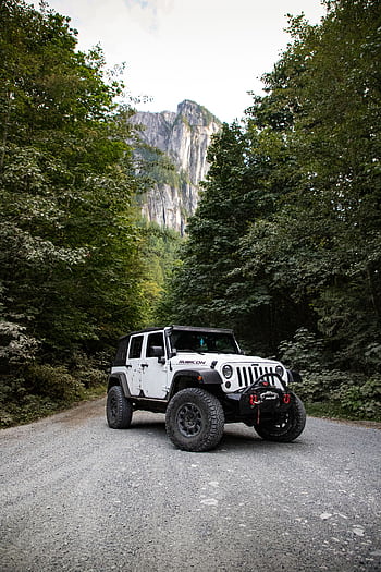 A rugged Jeep Wrangler on a gravel road surrounded by lush forest and towering mountains. HD phone wallpaper