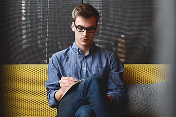 A young man in glasses writes in a notebook while sitting on a stylish couch indoors. HD wallpaper