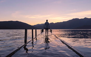 Man stands on a dock at sunset, reflecting on calm lake waters and mountainous backdrop in New Zealand. HD wallpaper